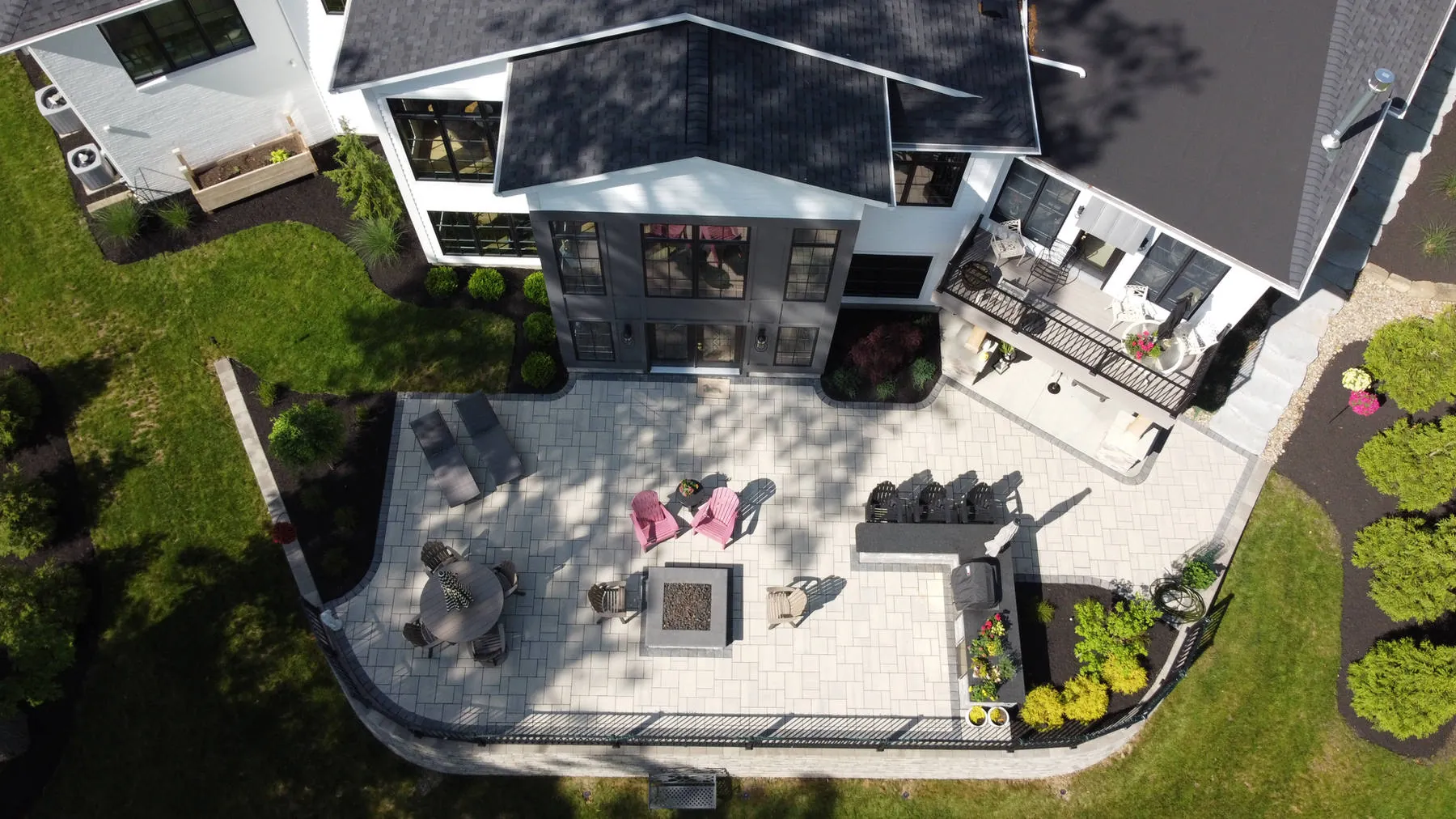 Overhead view of a large light-gray paver patio with a square fire pit, lounge seating, low edge walls, and planted beds behind a white house