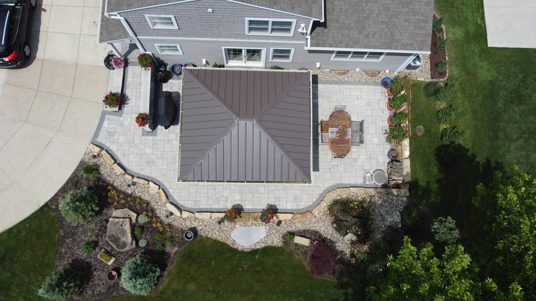 Overhead view of a metal-roofed pavilion on a paver patio, edged with natural wall stones, river rock, boulders, and flower beds