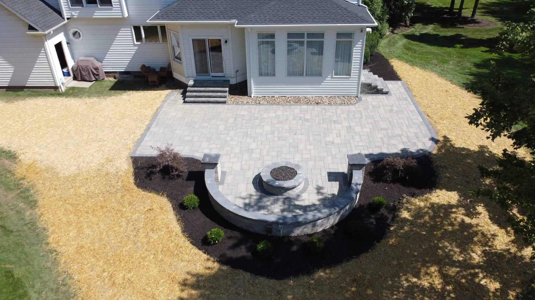 After: the same house with a new paver patio, a curved seat wall around a round fire pit, and new beds, with the lawn just seeded under straw