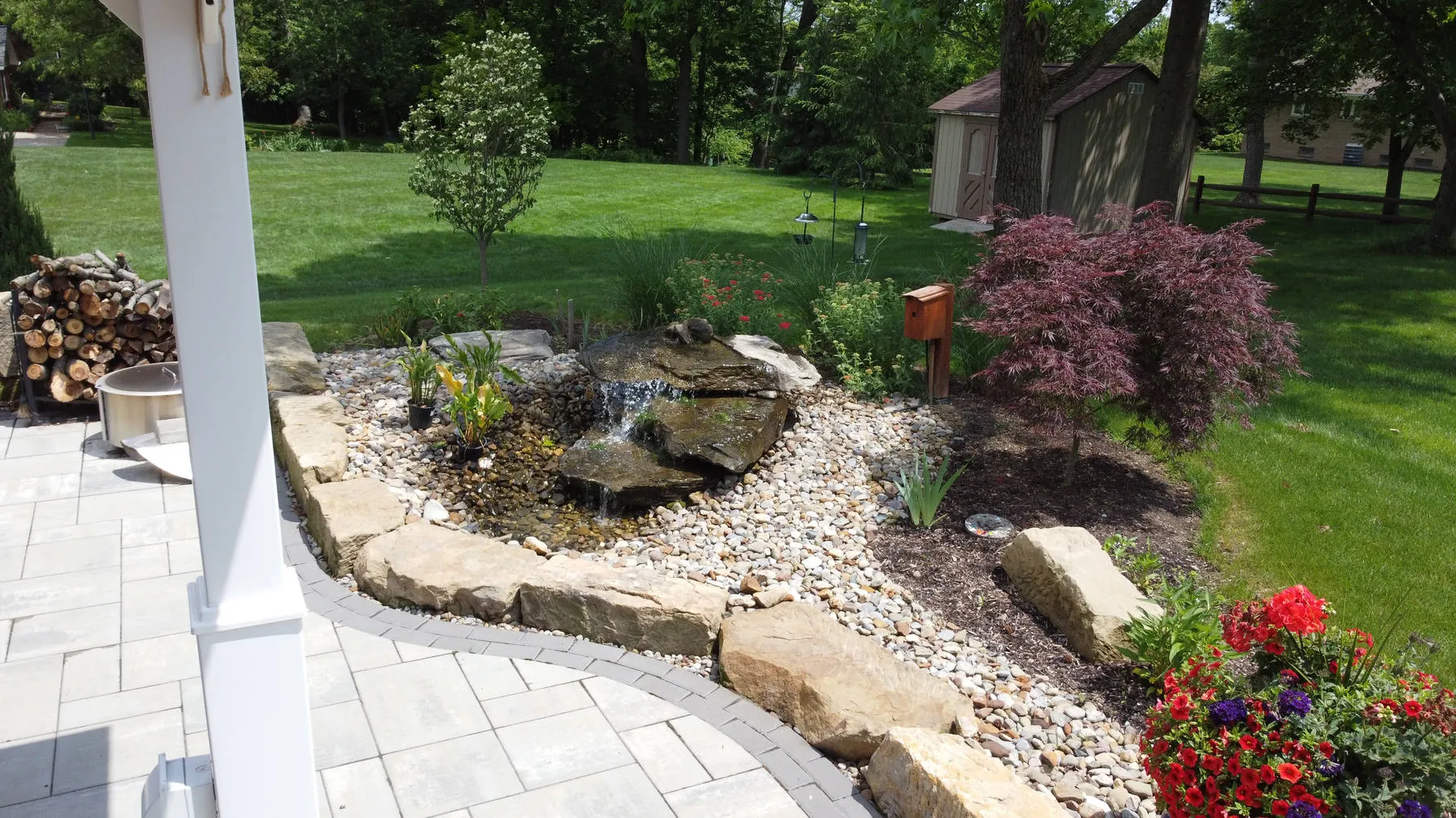 Boulder water feature spilling into a river rock bed beside a paver patio, with a Japanese maple, red flowers, and lawn