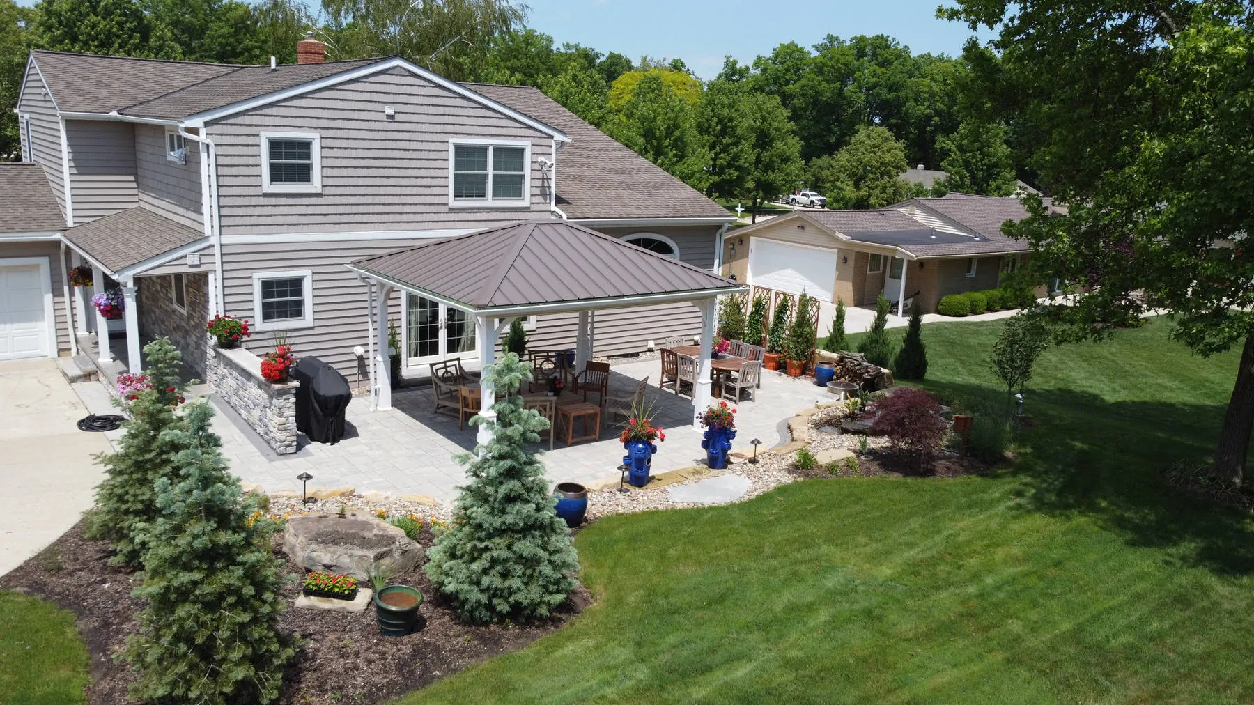Roofed pavilion over a dining table on a paver patio, with trellis panels, a boulder bed, a Japanese maple, and a wide lawn