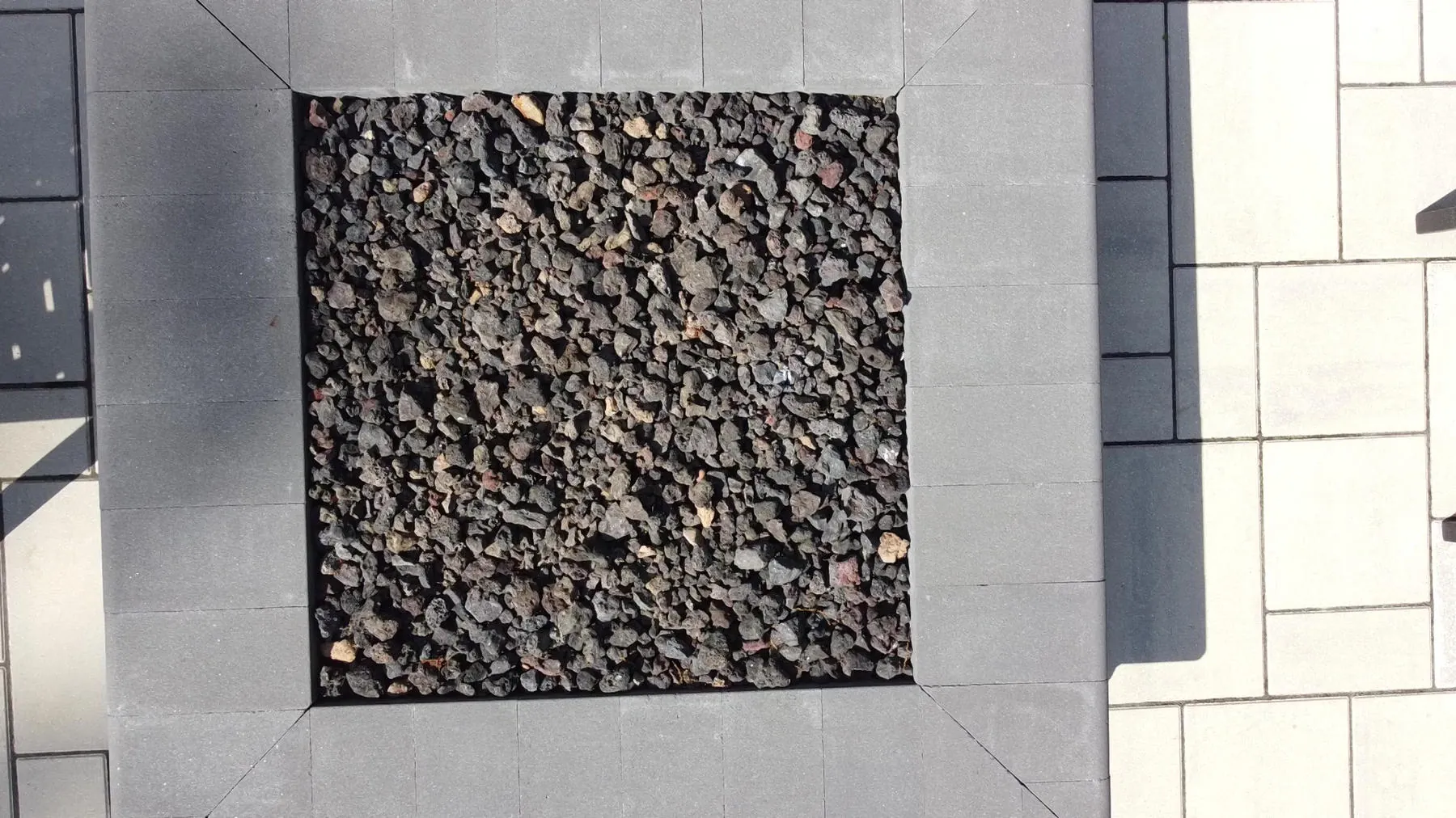Looking straight down into a square fire pit filled with lava rock, framed by smooth stone caps and large pavers
