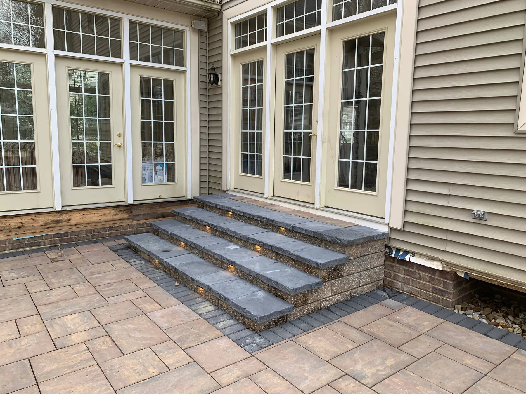 Three wide paver steps with built-in riser lights leading up to a row of French doors, on a tan paver patio