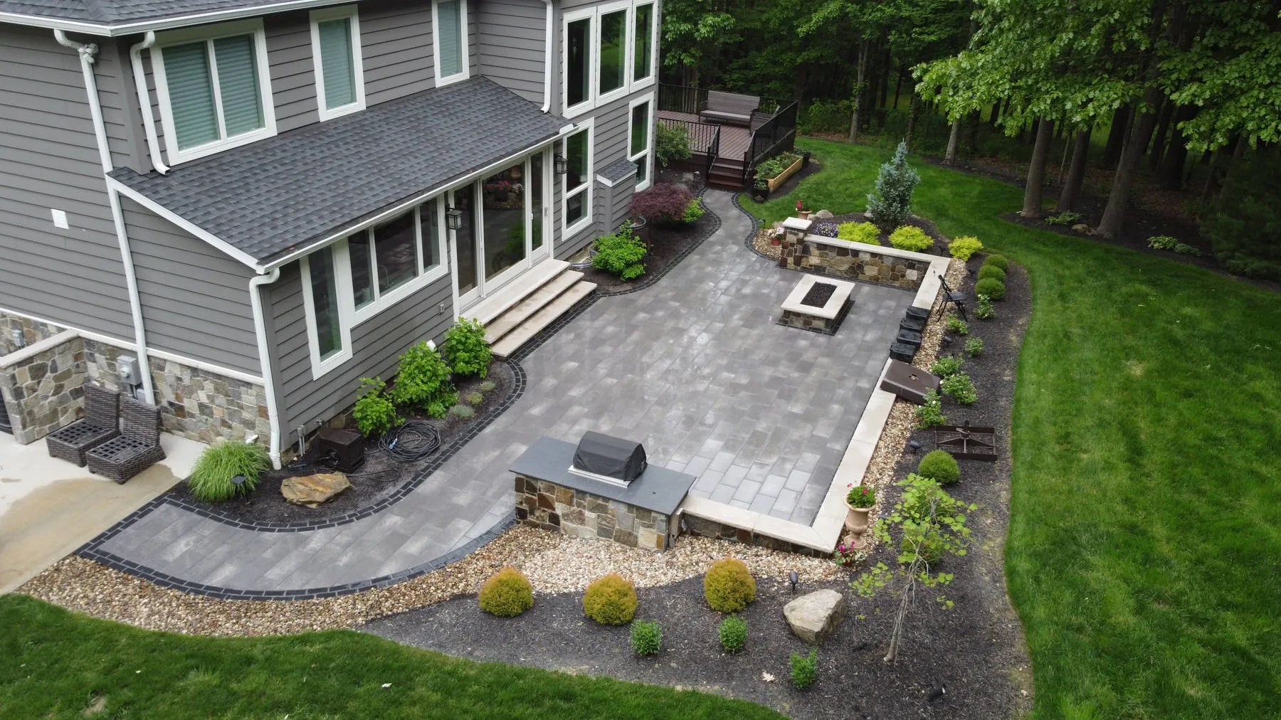 Wraparound paver patio with a stone-faced grill island, a square fire pit, and a raised stone wall bed beside a mowed lawn