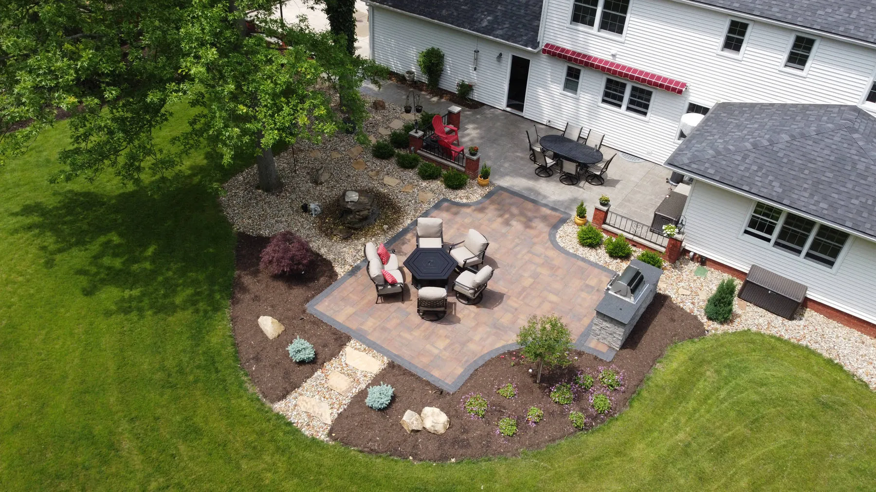 Aerial view of a banded paver patio with lounge chairs, a built-in grill station, a dining patio by the house, and curved planting beds edged in lawn