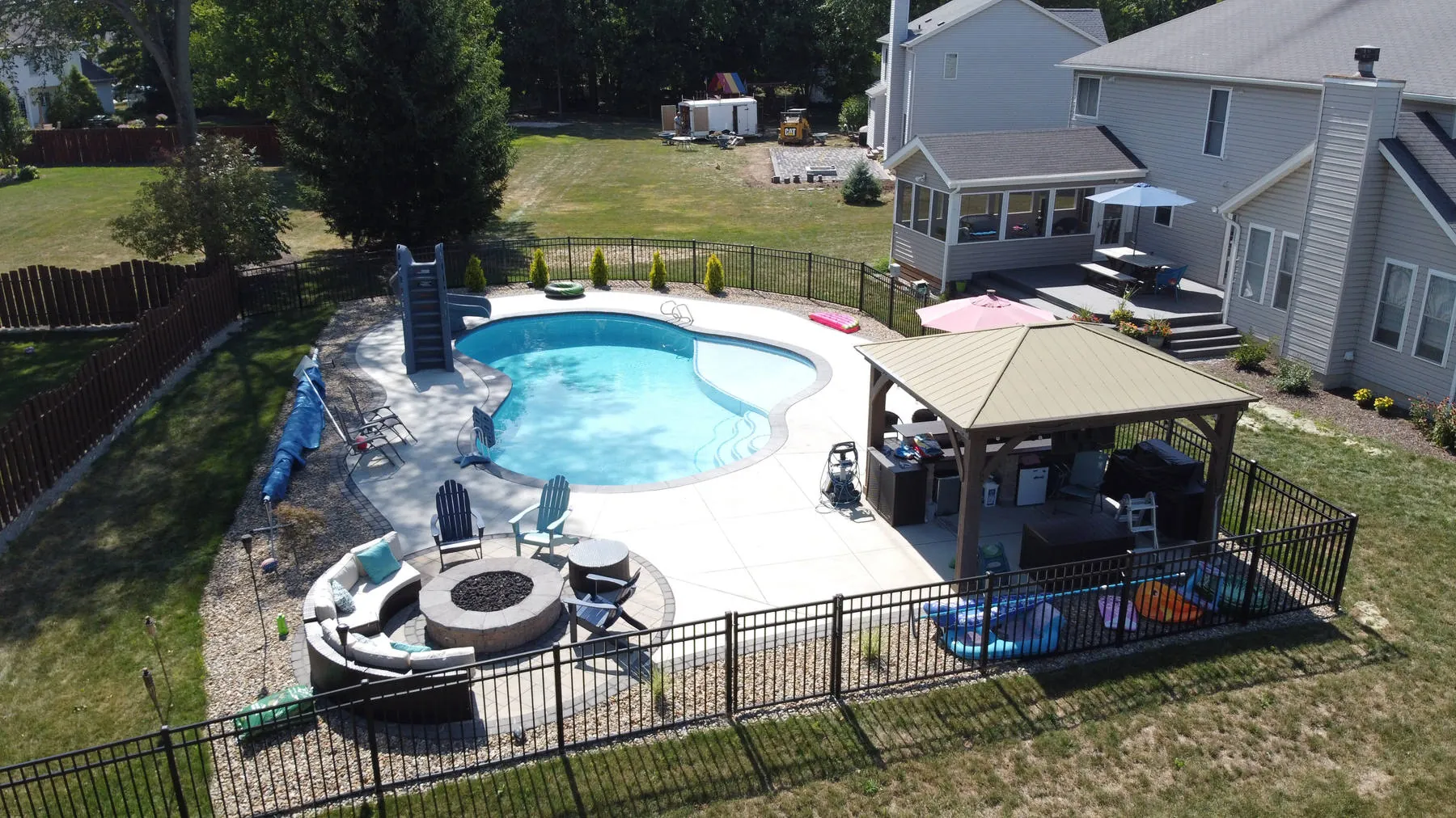 Pool surround with a roofed pavilion sheltering a counter and mini fridge, plus a round fire pit ringed by lounge seating inside the pool fence