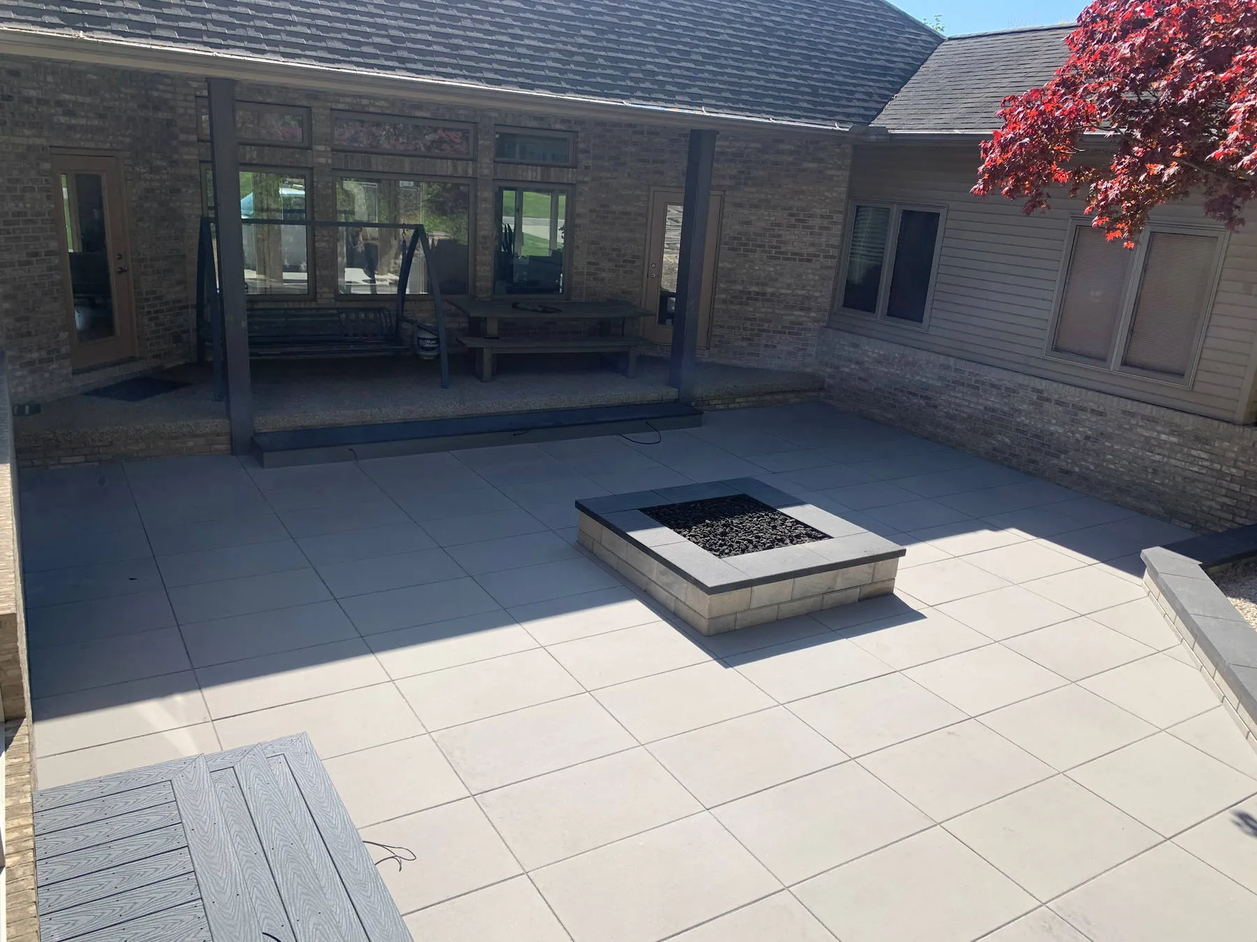 Square fire pit filled with lava rock on a large-format light-gray patio beside a covered porch