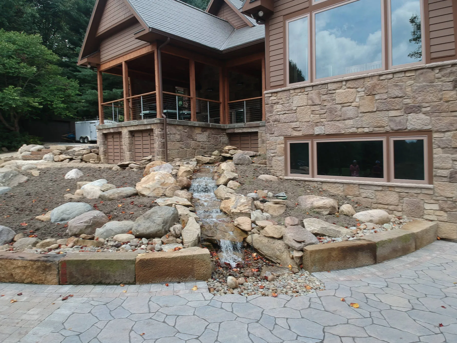 In progress: a boulder waterfall stepping down beside a stone-faced foundation to a flagstone-pattern patio, beds not yet planted