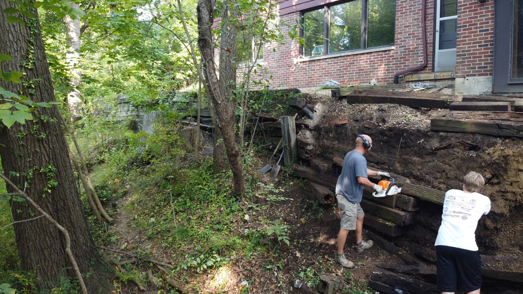 In progress: two crew members building a timber retaining wall on a wooded slope behind a brick house