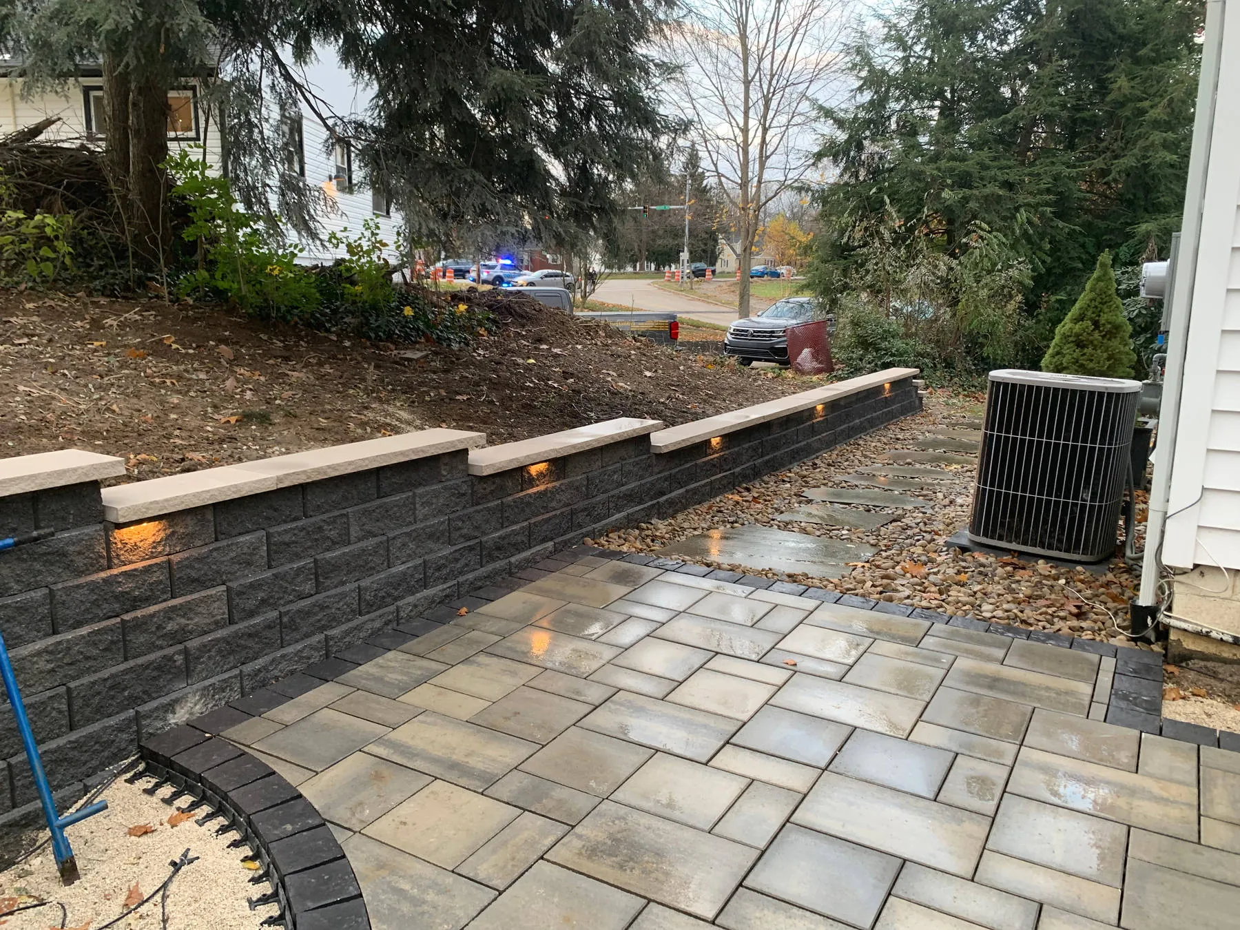 In progress: a block wall with lit capstones along a wet paver patio, with the slope above still bare