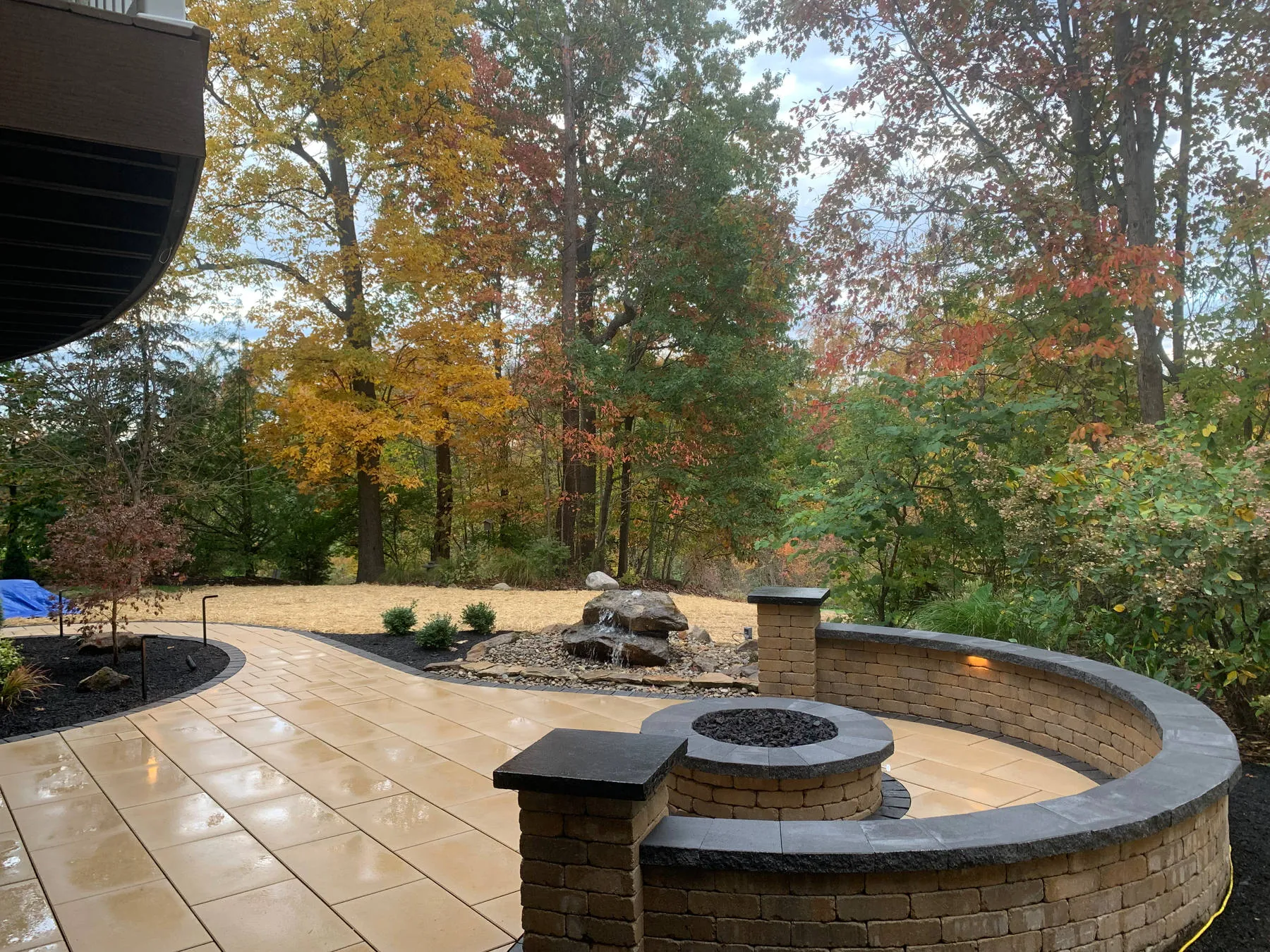 Just installed: a curved seat wall and fire pit on a tan paver patio with a small boulder bubbler behind, new lawn under straw and autumn trees