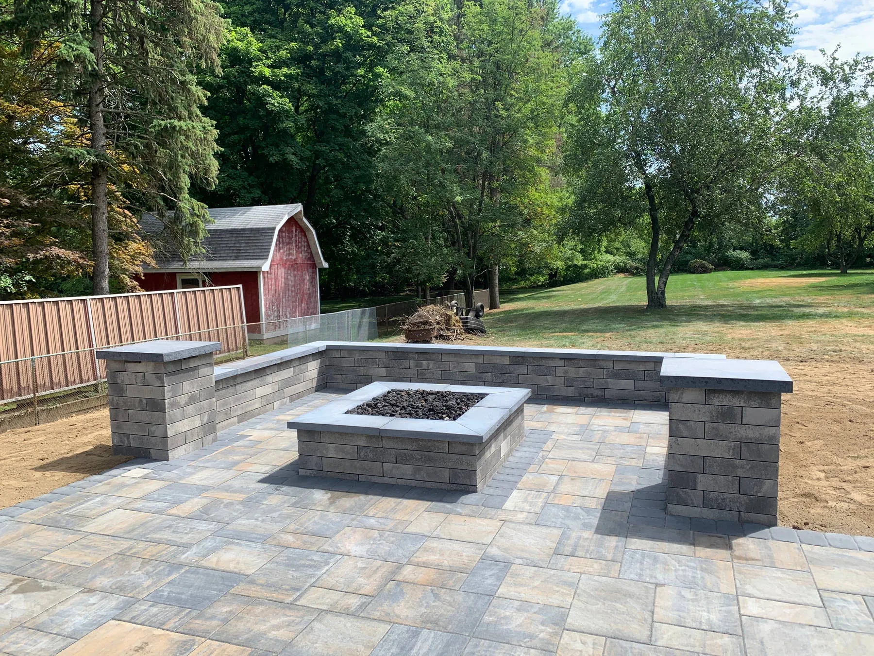 Just installed: a square fire pit and seat walls with pillars on a paver patio, edges still bare soil, red barn in the background