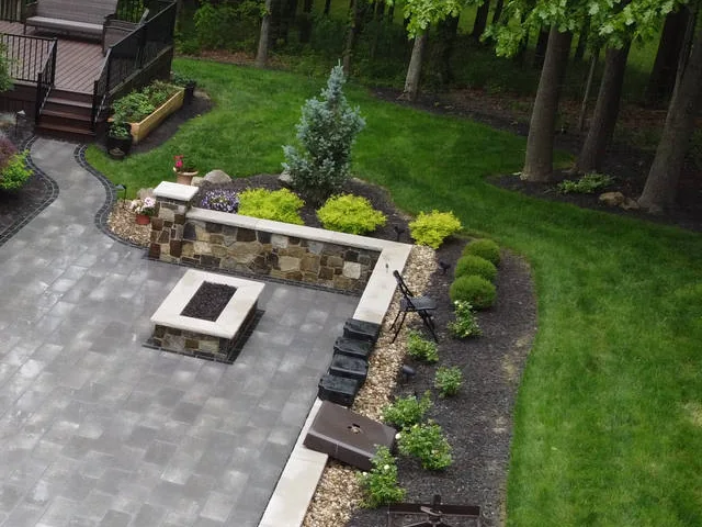 Stone-faced retaining wall with a light capstone holding up a raised planting bed beside a gray paver patio and square fire pit