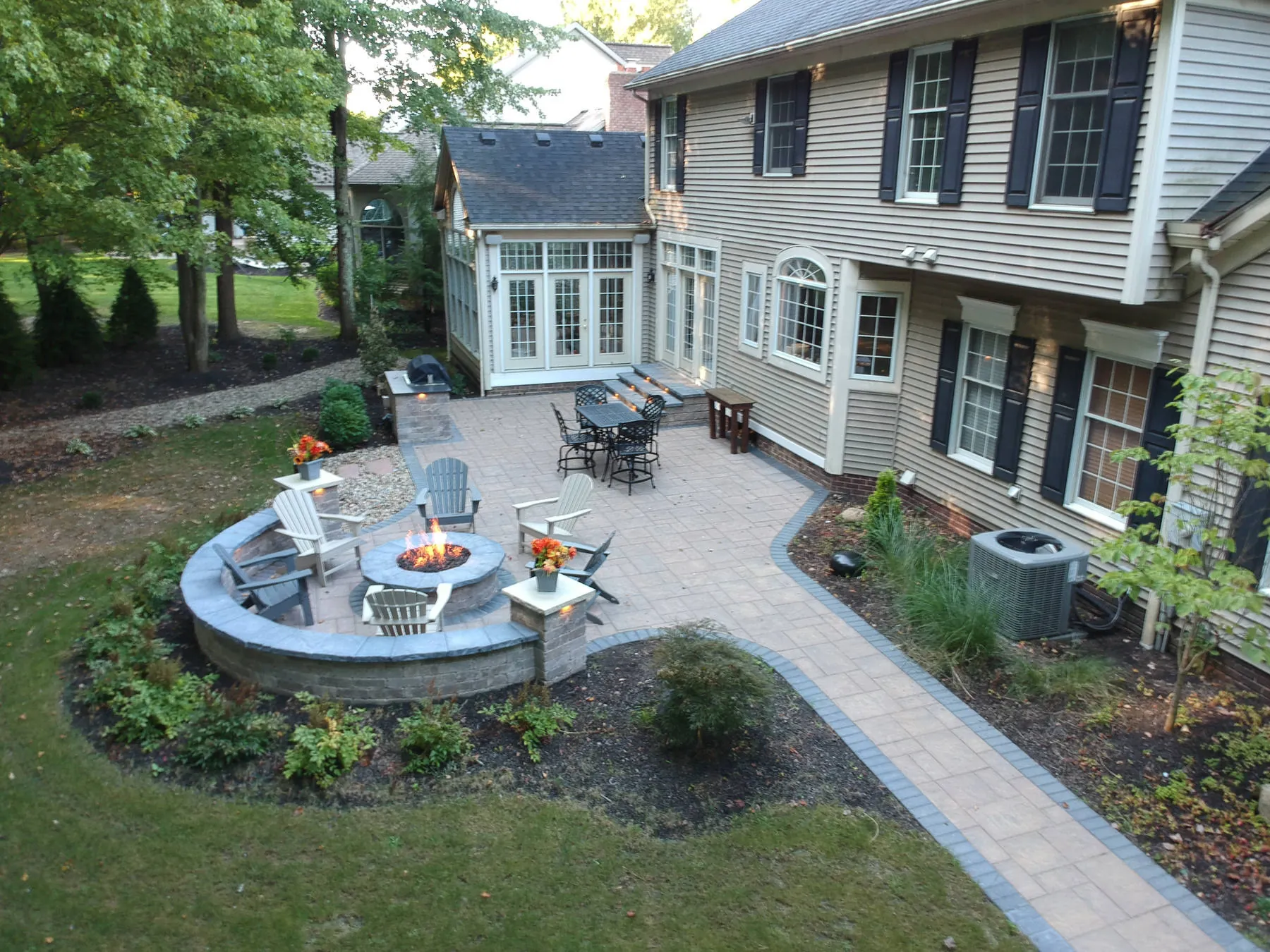 Curved block seat wall wrapped around a lit round fire pit, with a paver walkway and planted beds at dusk