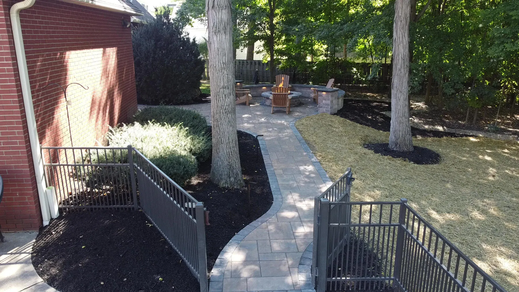 Paver walkway running between fresh mulch beds and shrubs along the side of a brick house, toward a patio with a fire pit